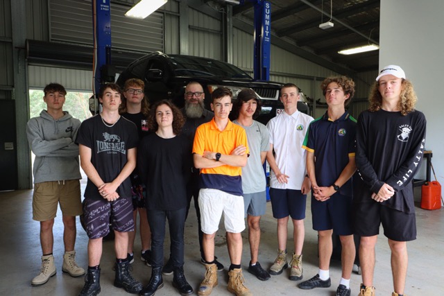 trainer and eight students standing in front of a car being jacked in the workshop